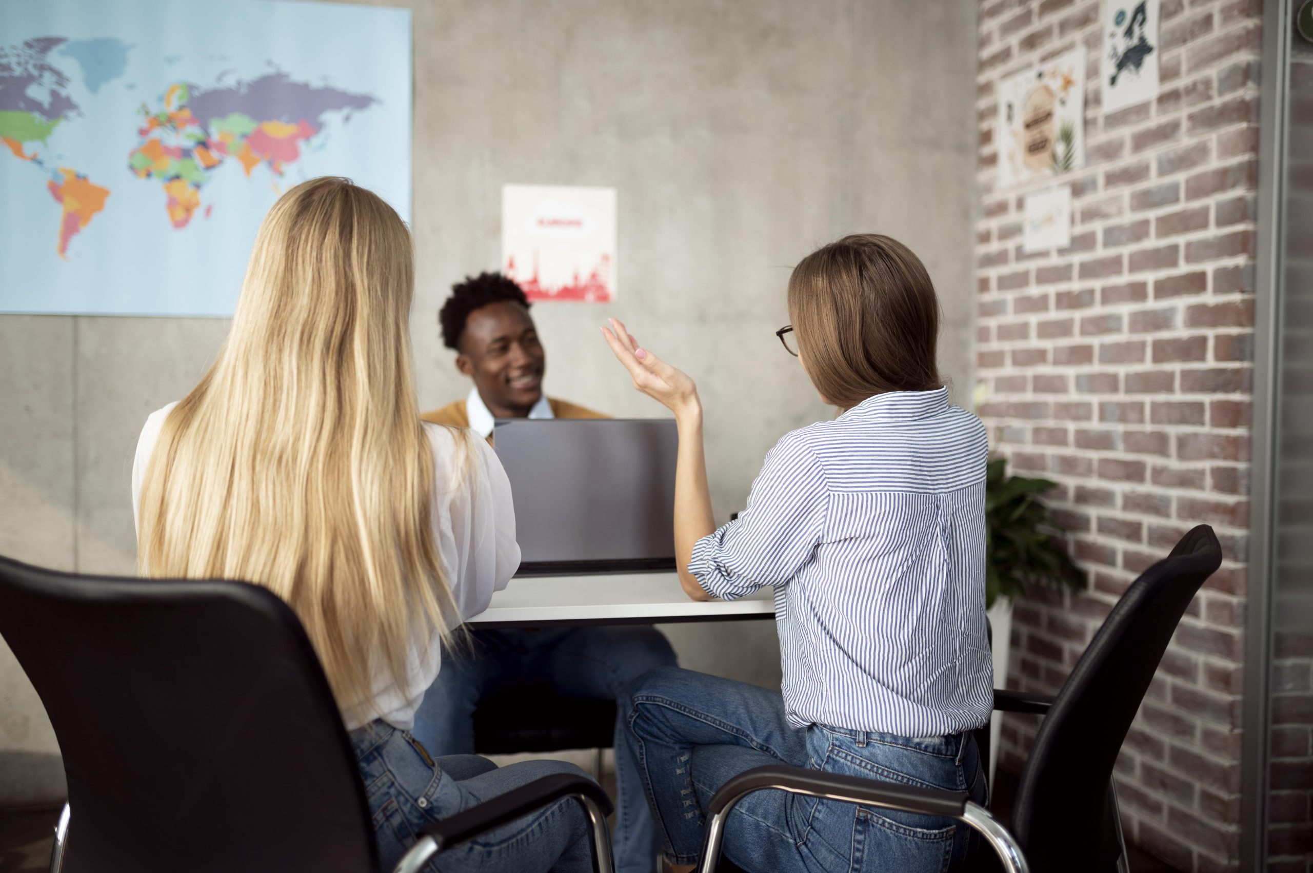 dos chicas sentadas una junto a otra, miran a un chico que habla con ellas, de fondo una oficina con un mapa del mundo en la pared