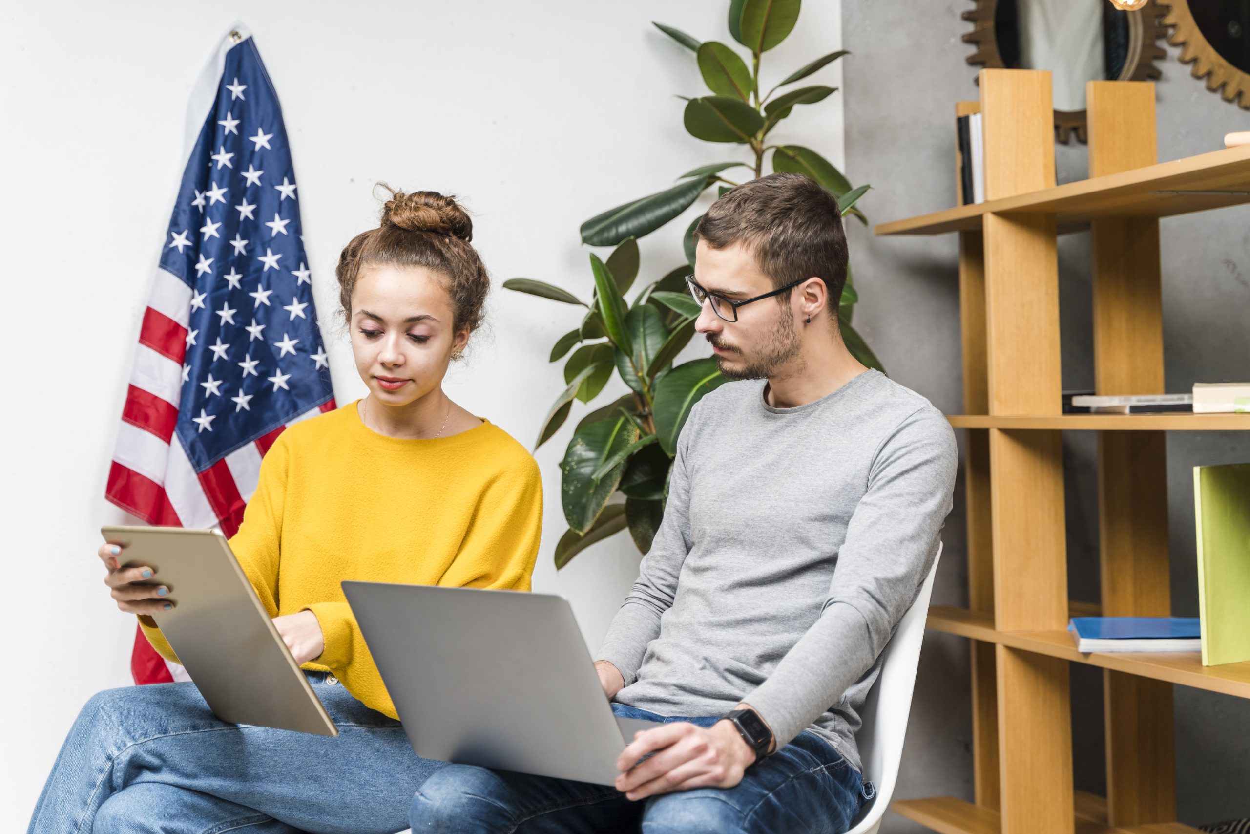 dos personas sentadas una junto la otra con un computador, de fondo la bandera de estados unidos