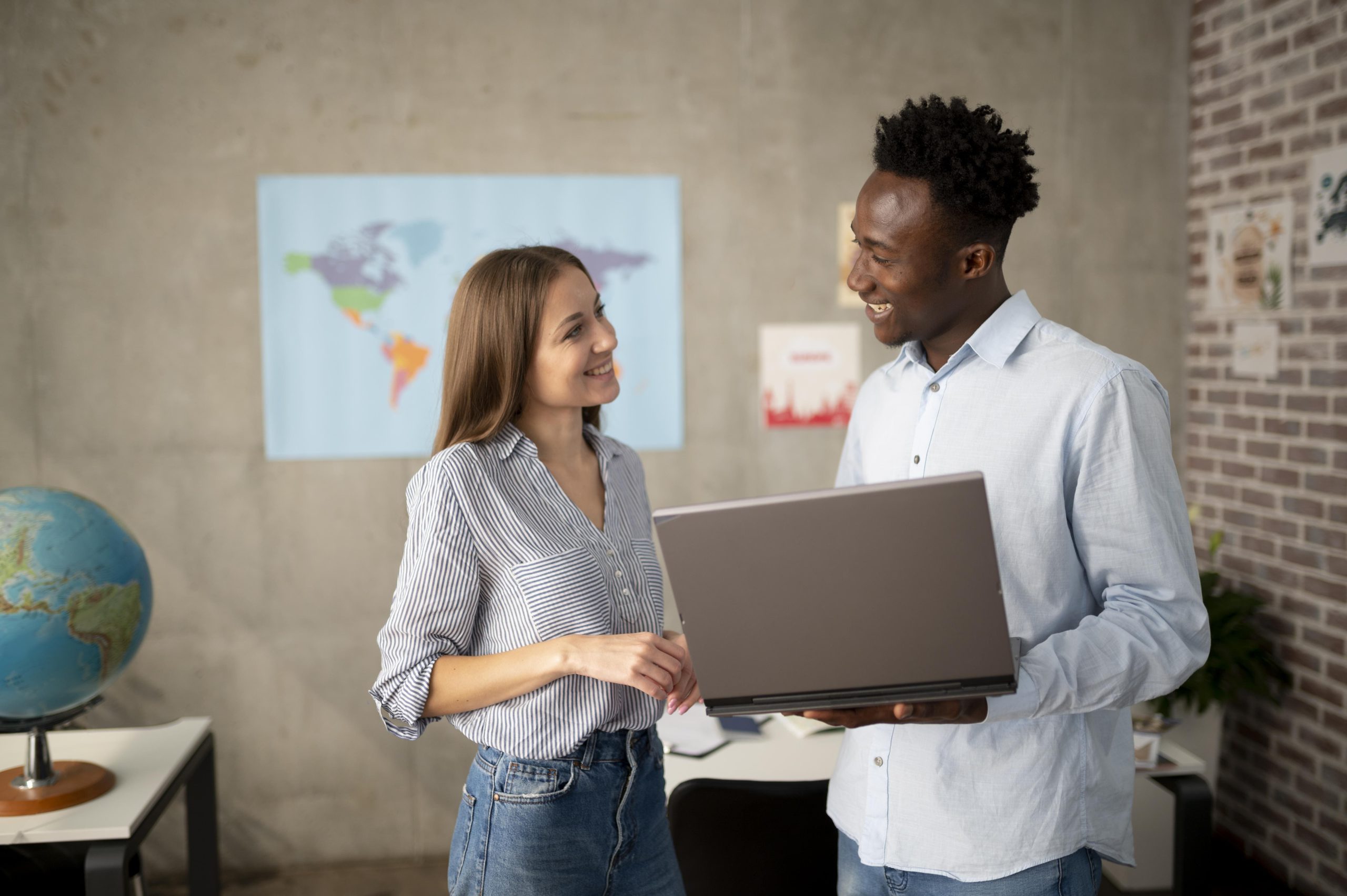 dos personas adultas una de ellas sostiene un computador portatil y ambas se miran a los ojos, de fondo una oficina con un mapa del mundo en la pared